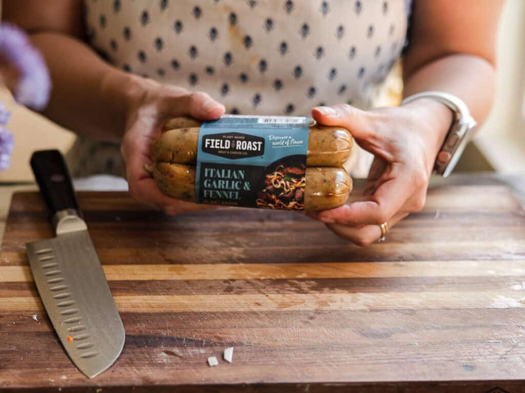 A person holds a package of Field Roast Italian Garlic & Fennel plant-based sausages. A knife is on a wooden cutting board in front of them. The setting appears to be a kitchen.